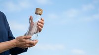 Person holding glass bottle with cork stopper against blue sky, suitable for beverage, eco-friendly product, sustainability, and wellness marketing campaigns.