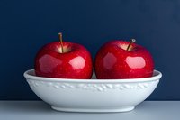 Two glossy red apples resting in a white ceramic bowl against a deep navy blue background, fresh healthy fruit still life.