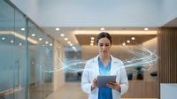 Female doctor in white coat using digital tablet with futuristic holographic data streams in modern hospital corridor for healthcare technology concepts.