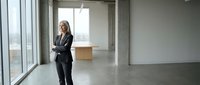 Businesswoman in formal black suit standing in empty modern office space with large windows and concrete flooring, suitable for corporate and professional business concepts.