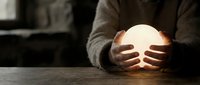 Businessman holding glowing orb of light in cupped hands at wooden desk, symbolizing innovation, creativity and future vision in corporate environment.