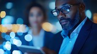 African American businessman wearing glasses reviews digital tablet in evening office setting with blue bokeh lights and colleague in background.
