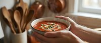 Young adult with light skin tone holding a bowl of tomato soup garnished with herbs and cream, styled kitchen setting with wooden utensils and natural window light for food photography and culinary content.