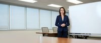 Professional businesswoman in navy suit standing confidently in modern conference room with large windows and whiteboard for corporate presentations.