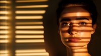 Young woman's face illuminated through window blinds creating dramatic shadow stripes in warm golden light, artistic portrait with moody atmospheric lighting.