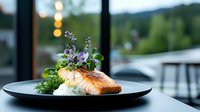 Grilled salmon fillet with fresh herbs and edible flowers served on black plate, garnished with microgreens and creamy sauce against blurred restaurant view.