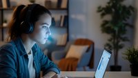 Young Hispanic woman wearing headphones working late on laptop computer in modern home office with warm lighting and plants in background.