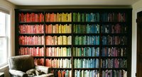 Home library with books organized by color creating rainbow spectrum on floor-to-ceiling shelves. Reading nook with leather chair for book lovers and bibliophiles.