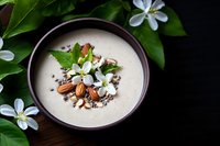 Creamy smoothie bowl topped with almonds, seeds and fresh white flowers, garnished with green leaves on dark background. Healthy breakfast concept.