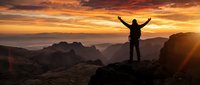 Silhouette of triumphant hiker with raised arms celebrating success on mountain peak at golden sunrise with dramatic orange sky and layered mountain ranges.