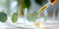 Golden essential oil drop falling from dropper into glass bottle, with fresh eucalyptus branch in soft focus background on white surface.