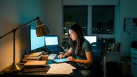 Young professional woman working late at night in modern home office with computer and desk lamp creating productive workspace atmosphere.