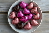 Heart-shaped chocolate candies wrapped in pink, magenta and brown metallic foil arranged on white ceramic plate against rustic wooden background.