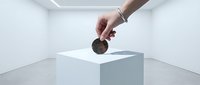 Hand dropping coin into white cube donation box in minimalist gallery space, symbolizing charity, investment, and financial contribution for fundraising campaigns.