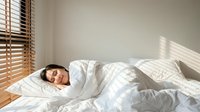Young woman peacefully sleeping in bright bedroom with natural sunlight streaming through wooden blinds onto white bedding for wellness content.