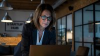 Mature woman with shoulder-length brown hair and glasses, wearing professional business attire, working on laptop in modern office space with industrial lighting and windows.