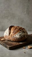Freshly baked artisan sourdough bread loaf on rustic wooden cutting board with flour dusting and crumbs scattered around against textured concrete background.