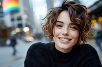 Young Caucasian woman with short curly brown hair wearing black sweater smiling outdoors in urban setting with blurred city background and rainbow flag.