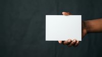 Person of African descent with medium-dark skin tone holding blank white card against dark textured background, suitable for mockups, advertising, and promotional campaigns.
