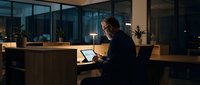 Mature man with gray hair working on laptop at wooden desk in modern office at night with city lights visible through large windows, suitable for business, technology, and corporate concepts.