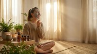 Young woman practicing mindfulness meditation with essential oils in sunlit room, sitting cross-legged on yoga mat surrounded by plants and aromatherapy bottles.