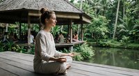 Young woman practicing meditation on wooden deck by tropical pond with traditional gazebo surrounded by lush green jungle vegetation for wellness retreat.