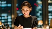 Young Asian woman smiling while writing in notebook at modern office desk with city lights background during evening work session.