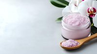 Pink sea salt in glass jar and wooden spoon with white orchid flower and green leaves on light background, spa wellness concept.
