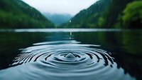 Raindrops create concentric ripples on calm mountain lake surface with misty forest hills in background, moody atmospheric nature scene in blue tones.