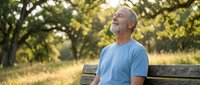 Mature man with gray hair and beard, Caucasian ethnicity, wearing light blue t-shirt, sitting on wooden bench in park surrounded by green trees and natural sunlight, contemplative expression, outdoor leisure and relaxation concept.