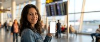 Young woman with curly brown hair holding smartphone with blank white screen at busy airport terminal, perfect mockup for travel apps and mobile advertising.