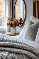 Cozy bedroom interior with chunky knit blanket, soft pillows, oval mirror, and dried flowers in white vase. Natural morning light creates warm atmosphere.