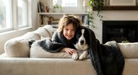 Young girl with brown hair cuddling black and white border collie dog on beige sofa in cozy living room with fireplace and bookshelves in background.