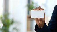 Mature African American professional in formal business attire holding blank white card at chest level in modern office environment with blurred green plants and colleagues in background.