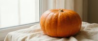 Large orange pumpkin resting on linen fabric near a bright window, evoking autumn harvest atmosphere for seasonal and holiday designs.