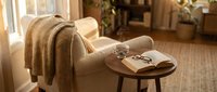 Cozy beige armchair with knitted throw blanket and open book on wooden side table in warm sunlit living room interior for home decor and relaxation.
