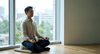 Young Asian man practicing meditation in lotus position by large window in modern office space with city view for mindfulness and wellness concepts.