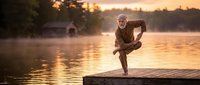 Young African American man practicing yoga crow pose on wooden dock at sunset over calm lake water with golden light reflection and peaceful atmosphere.