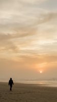 Solitary figure walking on empty beach during golden hour sunset with dramatic cloudy sky and calm ocean waves creating peaceful seascape atmosphere.