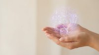 Human hand holding glowing purple energy sphere with swirling patterns against soft beige background for wellness and spiritual healing concepts.