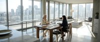 Two professional businesswomen having meeting in modern office conference room with city skyline view through floor-to-ceiling windows during daytime.