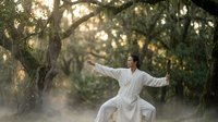 Asian woman practicing tai chi in peaceful forest setting with golden sunlight filtering through trees creating serene morning meditation atmosphere.