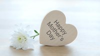 Wooden heart-shaped decoration with Happy Mother's Day text next to white chrysanthemum flower on light background for greeting card design.