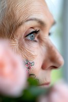 Close-up profile view of mature woman with blue eyes, facial tattoo and natural aging skin texture surrounded by soft pink floral elements in foreground.