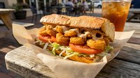 Crispy fried shrimp po'boy sandwich with lettuce and tomato on crusty bread served with iced tea on outdoor wooden table for casual dining experience.