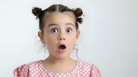 Surprised young Caucasian girl with bun pigtails and open mouth expressing shock and amazement on light background.