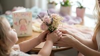 Mother and daughter hands holding small flower bouquet during Mother's Day celebration with greeting cards and gifts on table.