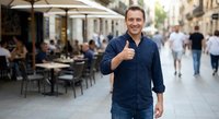 Middle-aged Caucasian businessman giving thumbs up gesture while walking on European city street with outdoor cafe terrace in background during daytime.