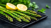 Fresh green asparagus spears on black grill pan with lemon halves, dark moody background. Healthy seasonal vegetable cooking concept for food photography.