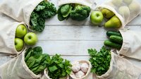 Fresh green vegetables and fruits arranged in circle on white wooden background with eco-friendly reusable bags for healthy eating and sustainable lifestyle.